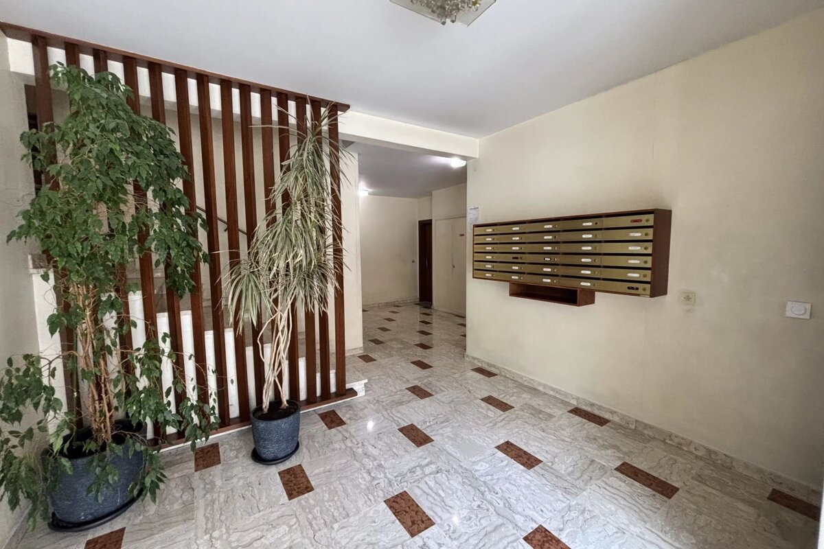 A building lobby with a row of mailboxes on a light wall, two large potted plants by a wooden divider, and a patterned tiled floor.