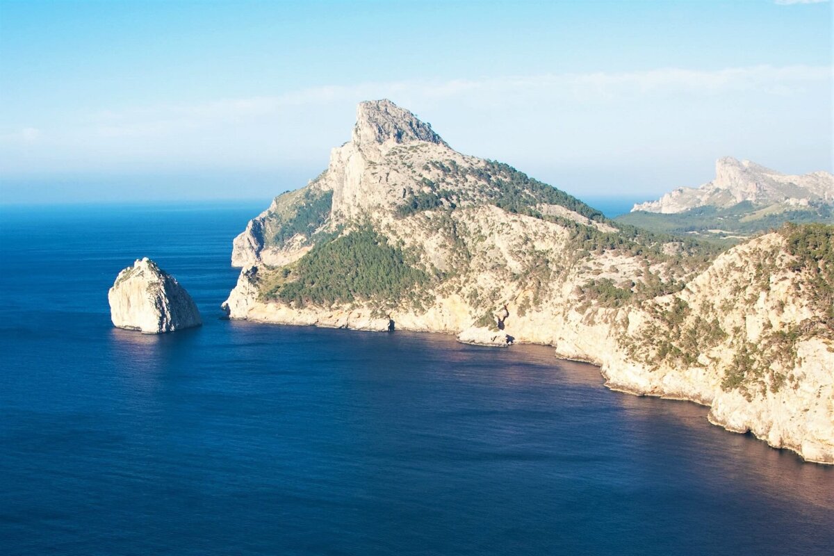 A stunning coastal view with deep blue sea meeting rugged, tree-covered cliffs and a prominent sharp peak. An isolated rock stands in the clear water.
