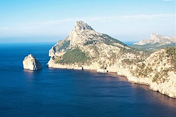 A stunning coastal view with deep blue sea meeting rugged, tree-covered cliffs and a prominent sharp peak. An isolated rock stands in the clear water.