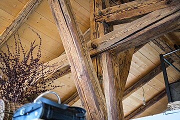 Rustic interior with prominent, dark wooden beams and a light wooden plank ceiling. Dried foliage and a blurred bag add to the cozy, natural ambiance.