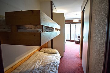 A room with wooden bunk beds and a single bed, next to a red-carpeted hallway leading to a bright room with a balcony.