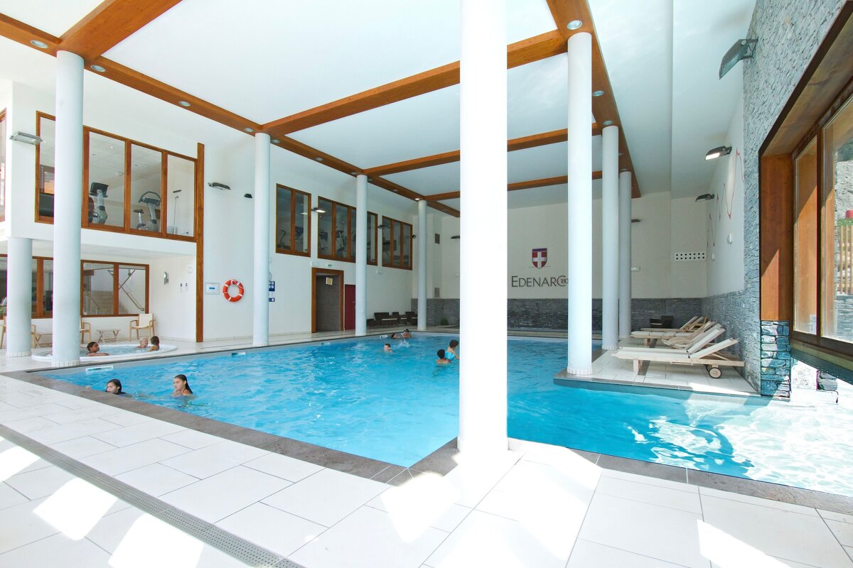 A spacious, bright indoor pool area with several people swimming and relaxing in a jacuzzi. A gym is visible upstairs through windows.