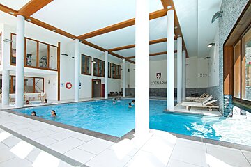 A spacious, bright indoor pool area with several people swimming and relaxing in a jacuzzi. A gym is visible upstairs through windows.