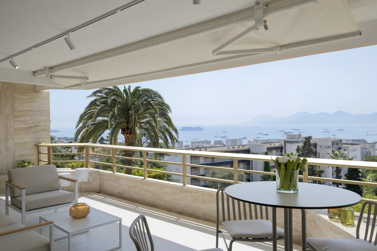 Modern balcony with stylish furniture under an awning, a palm tree, and a panoramic sea view over a city and distant mountains.