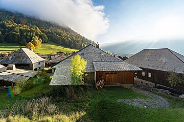 Alpine village homes with slate roofs, set against a vibrant autumn forest and misty mountains. Sun rays break through clouds.