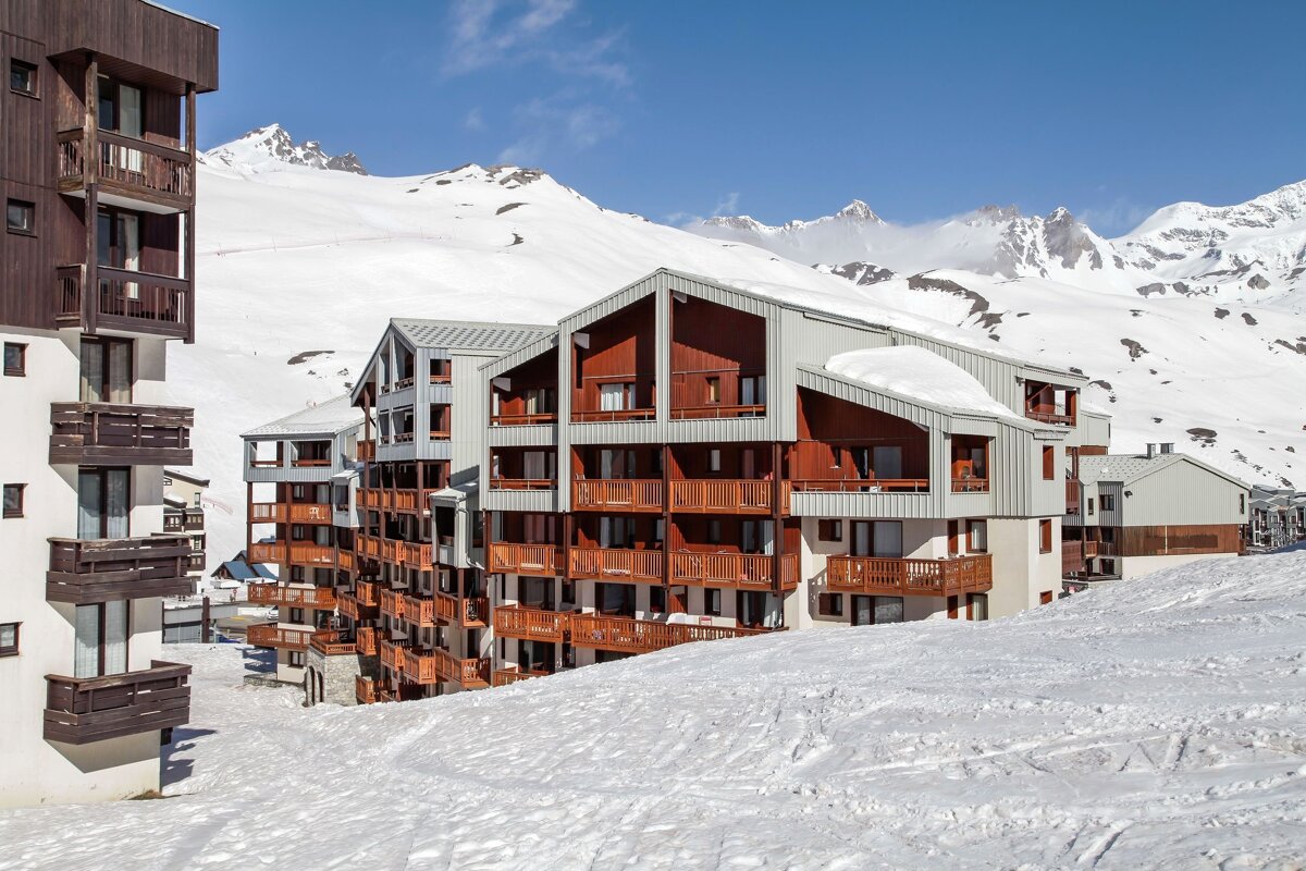 Alpine ski apartments with numerous balconies nestled in a snow-covered mountain landscape under a clear blue sky.