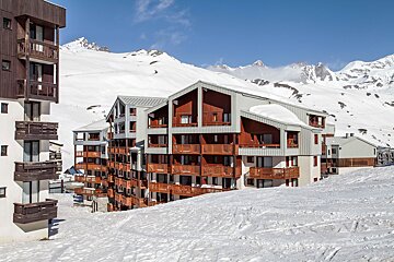 Alpine ski apartments with numerous balconies nestled in a snow-covered mountain landscape under a clear blue sky.
