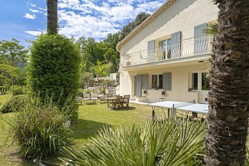 A Mediterranean-style villa with a lush green garden, patio furniture, and a ping-pong table under a bright, sunny sky.