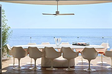 A modern outdoor dining area with white chairs and table on a wooden deck, boasting a vast blue ocean view under a clear sky. A ceiling fan hangs above.