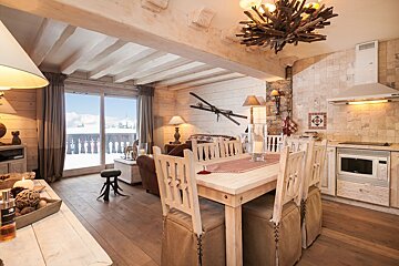 Rustic ski chalet interior with wood walls, beams, and an open layout encompassing living, dining, and kitchen areas. A snowy mountain view outside.