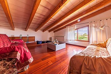 A bright attic living room featuring exposed wooden beams, polished hardwood floors, two cozy sofas, and a window overlooking lush greenery.