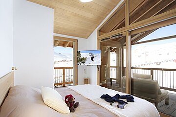Cozy bedroom with wooden ceiling, featuring a TV showing a skier, a comfortable bed, and a balcony overlooking stunning snowy mountains.