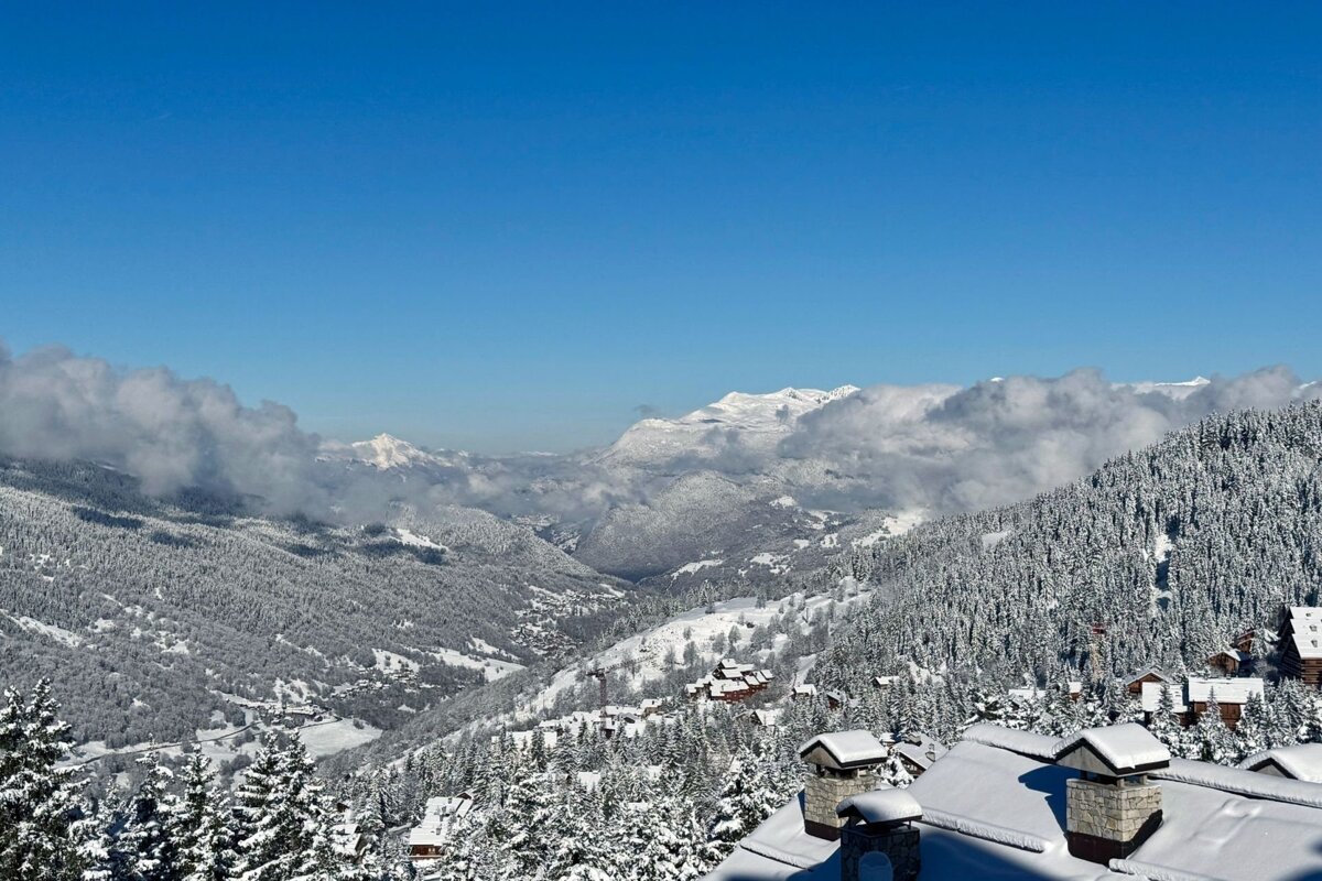 A vast snowy mountain landscape with frosted forests, a valley village, distant peaks under a clear blue sky with some clouds, and chalets.