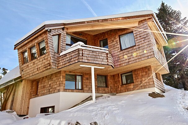 A wooden chalet with shingle siding sits on a snowy hillside under a clear blue sky.