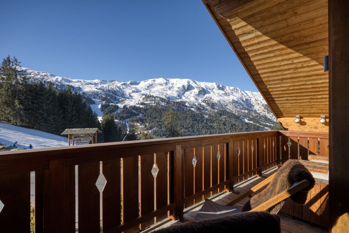Wooden balcony overlooking sunlit, snow-capped mountains, ski slopes, and pine forests under a clear blue sky.