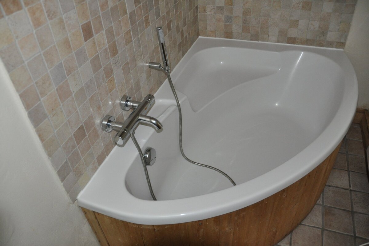 A modern white corner bathtub with chrome fixtures, set against a mosaic tiled wall and featuring wood paneling on its exterior.