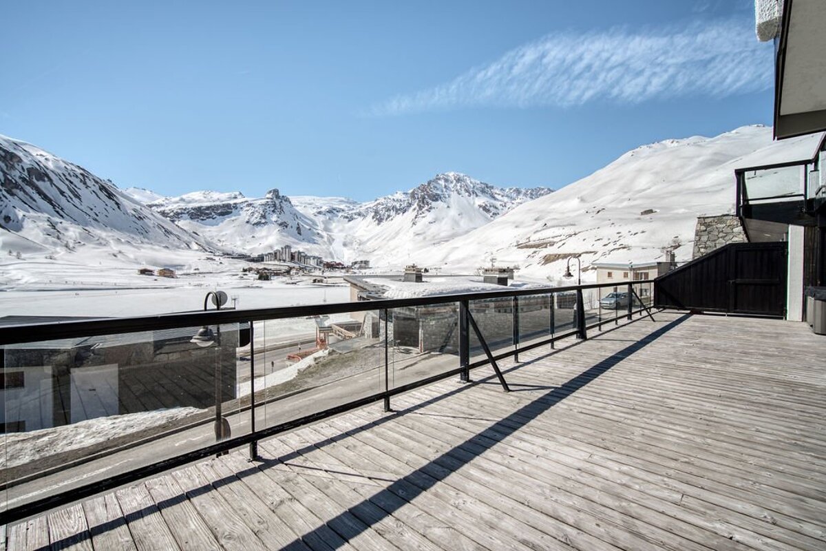Wooden deck balcony with a glass railing overlooking snow-covered mountains and a valley resort under a clear blue sky.