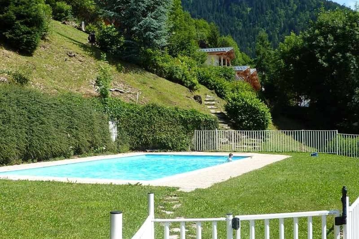 An outdoor swimming pool surrounded by green grass, with a forested hillside and chalets in the background.