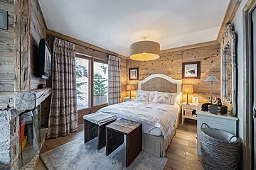 Rustic wooden bedroom with a cozy bed, fireplace, and large window showcasing a snowy mountain view. Warm and inviting chalet atmosphere.
