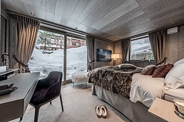 A cozy, luxurious bedroom with wood paneling, a fur throw on the bed, and large windows revealing a beautiful snowy winter scene outside.