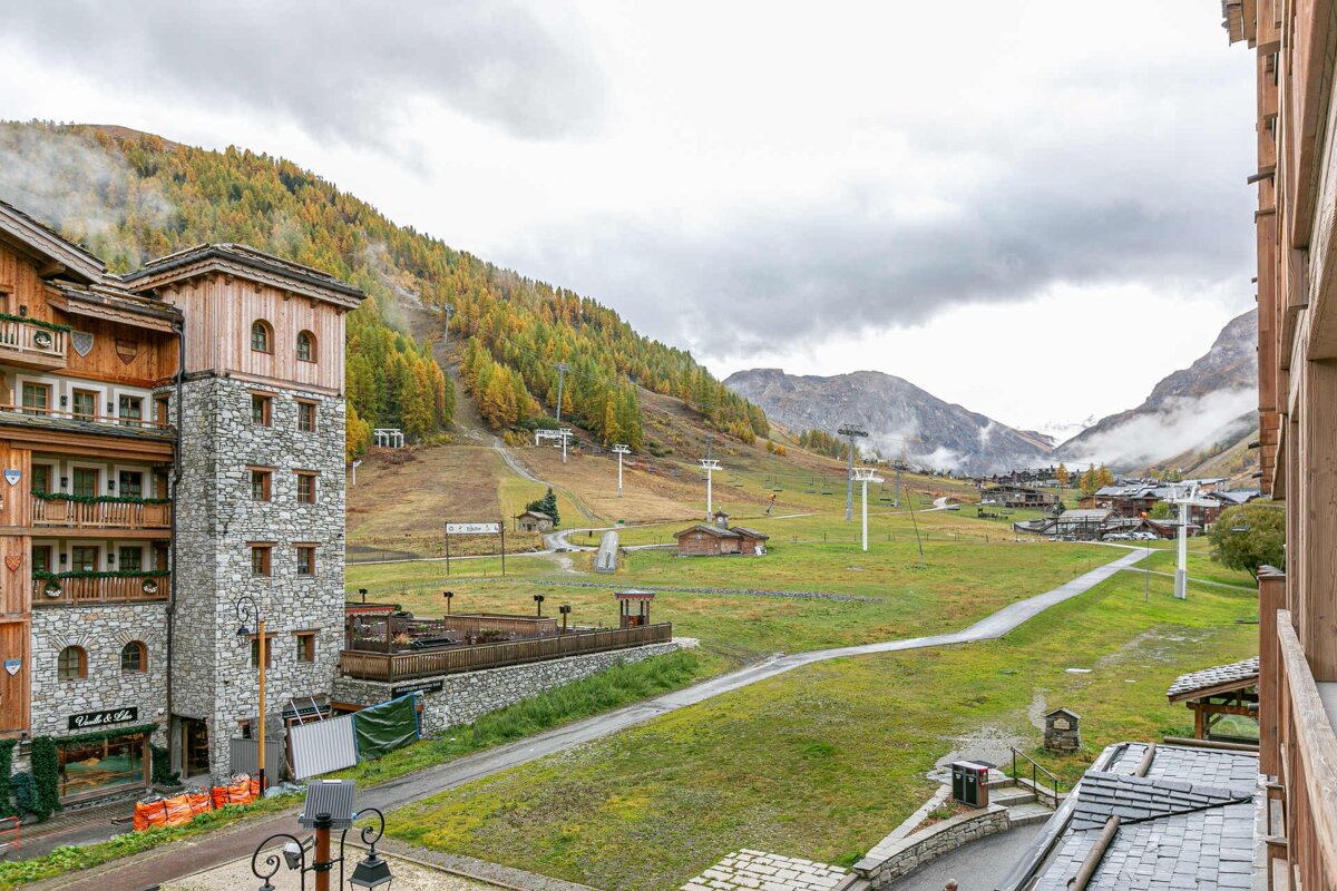 Autumn mountain resort view: traditional buildings, golden trees on ski slopes with lifts, green fields, and a cloudy sky.