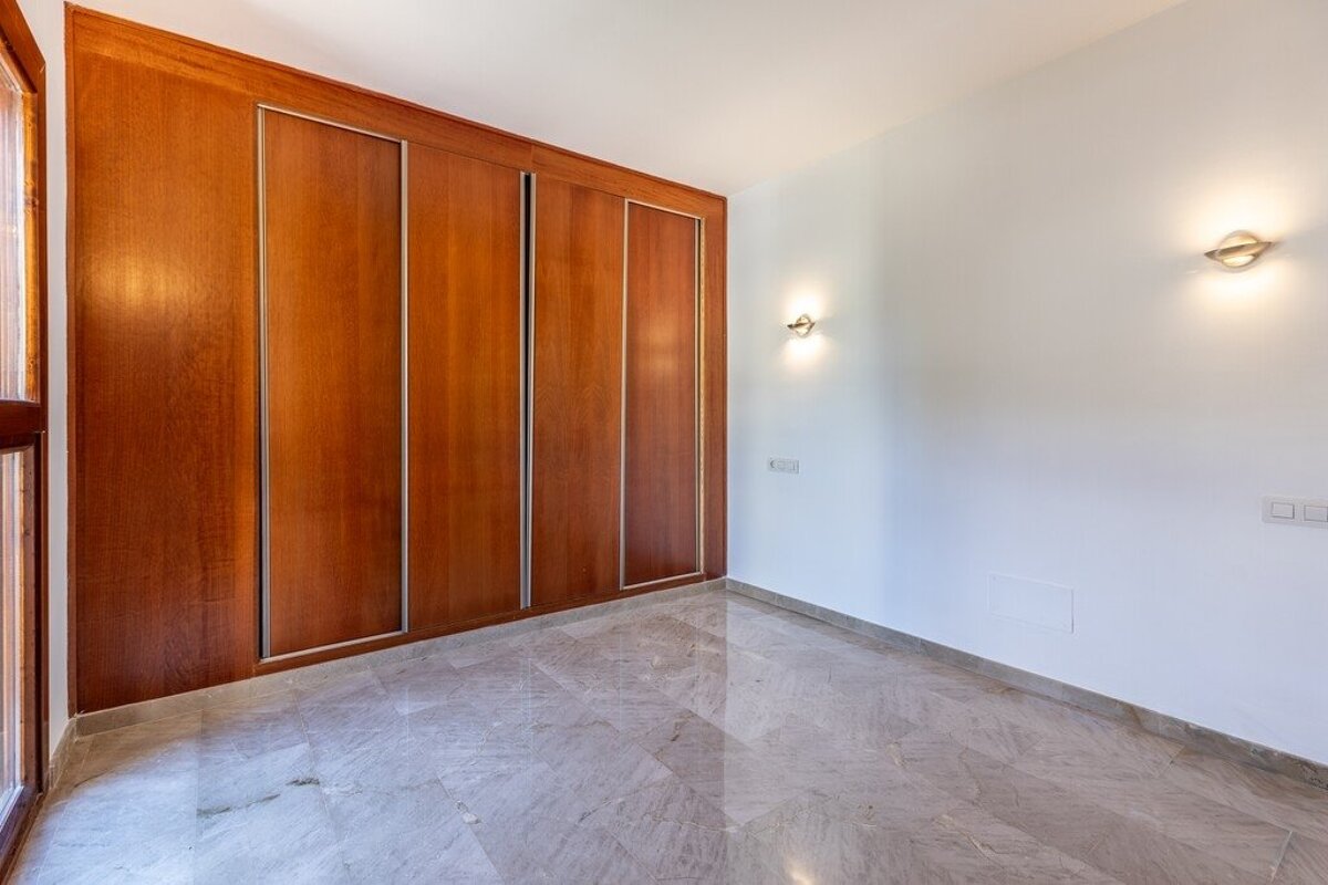 Empty room with a full-wall wooden wardrobe, white walls, two illuminated wall sconces, a marble floor, and a window.