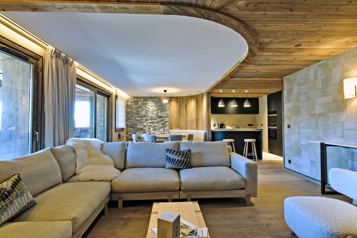 A modern, open-plan living room, dining area, and kitchen with wood ceilings, stone walls, a large sofa, and floor-to-ceiling windows.