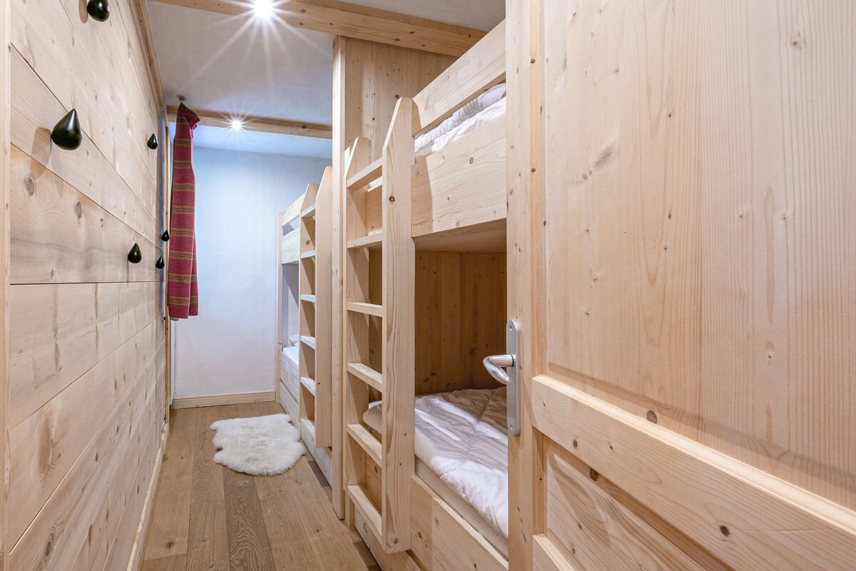 Cozy, light wood cabin-style hallway with built-in bunk beds, white bedding, dark wall hooks, and bright ceiling lights, creating a warm, rustic feel.