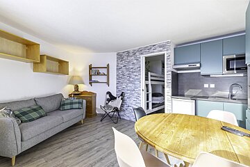 A modern apartment features a grey sofa, wooden dining table, and a blue kitchenette. Bunk beds are visible through an open doorway in a tiled wall.