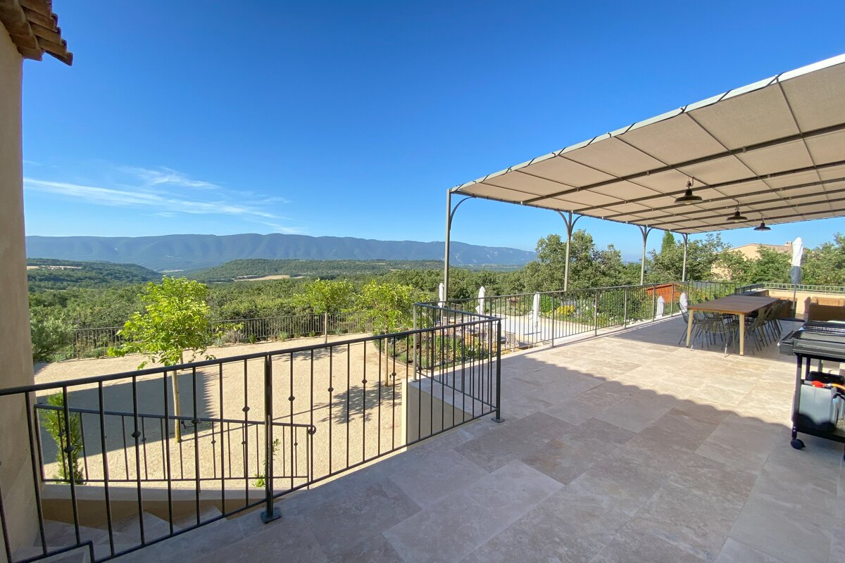 A sunny outdoor patio with a covered dining area, offering a panoramic view of a lush green valley and distant mountains under a clear blue sky.