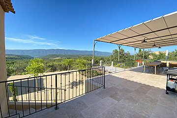 A sunny outdoor patio with a covered dining area, offering a panoramic view of a lush green valley and distant mountains under a clear blue sky.