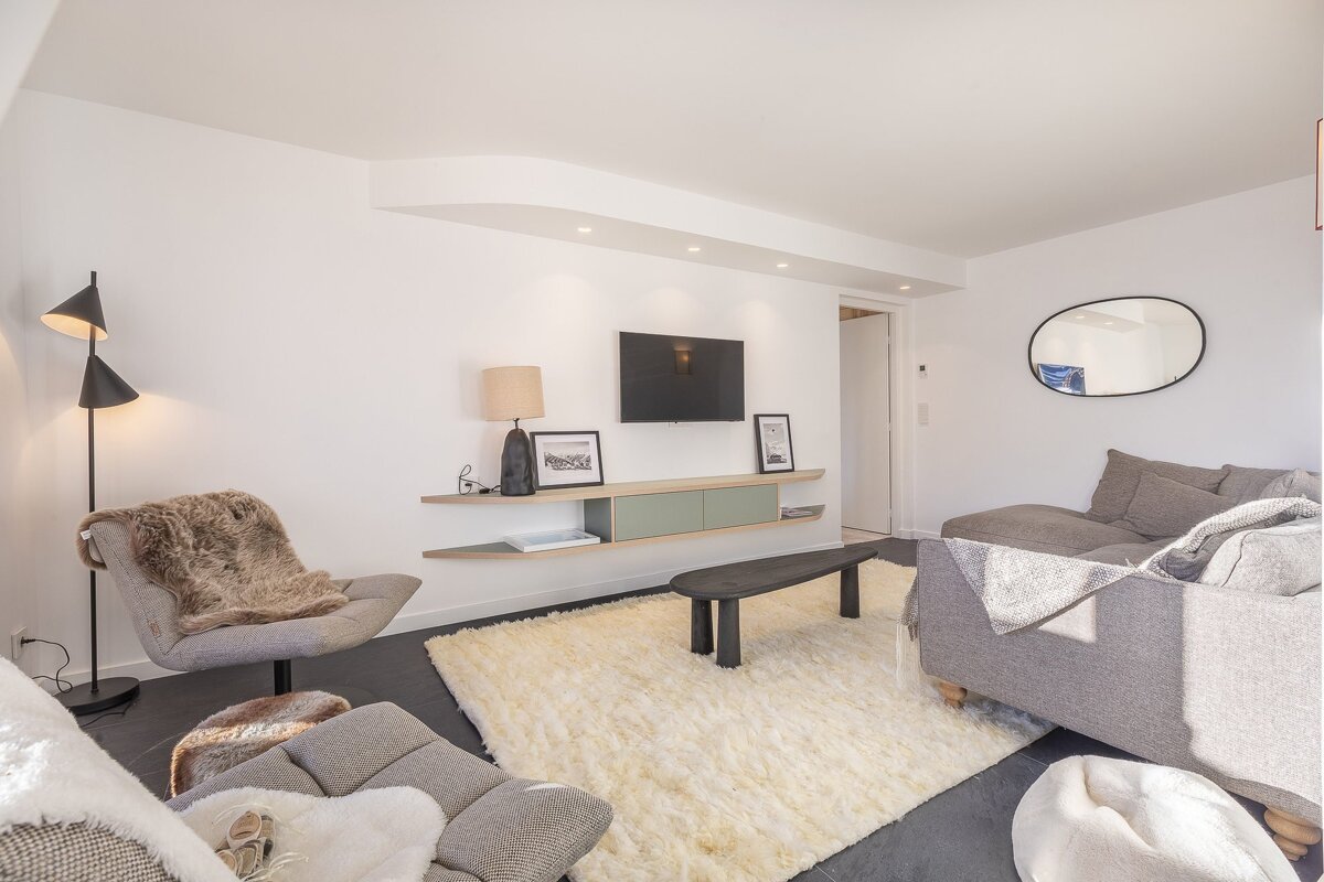 A cozy, modern living room with grey armchairs, a sofa, a fluffy white rug, a wall-mounted TV, and minimalist decor.