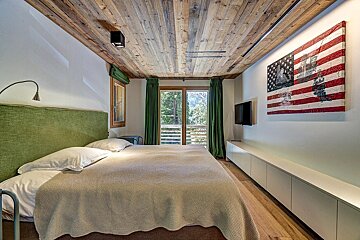 A cozy bedroom with a wooden ceiling, green accents, a balcony offering mountain views, and a large American flag artwork above a TV.