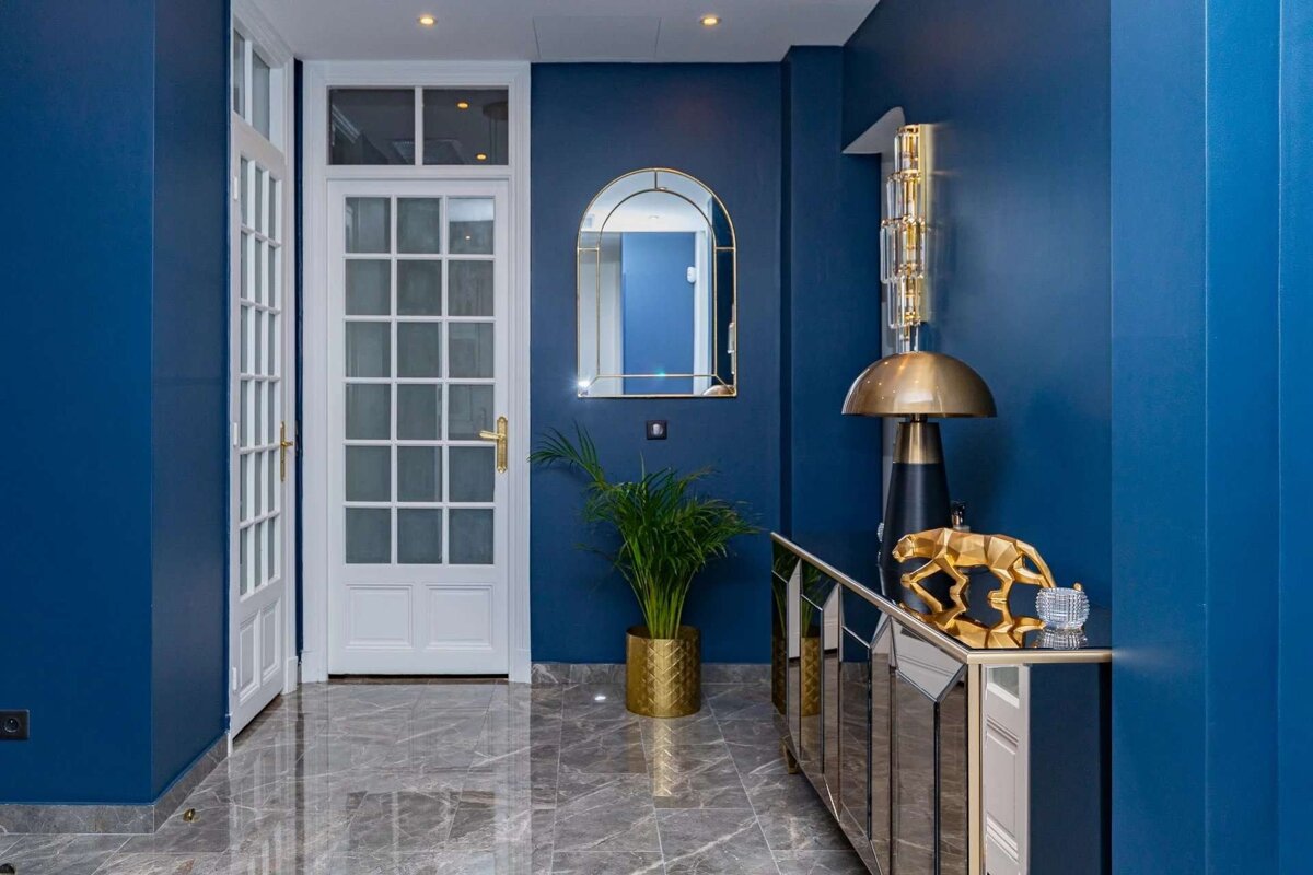 A modern blue hallway features white French doors, glossy marble floors, a gold arched mirror, elegant lamp, and a golden panther sculpture.