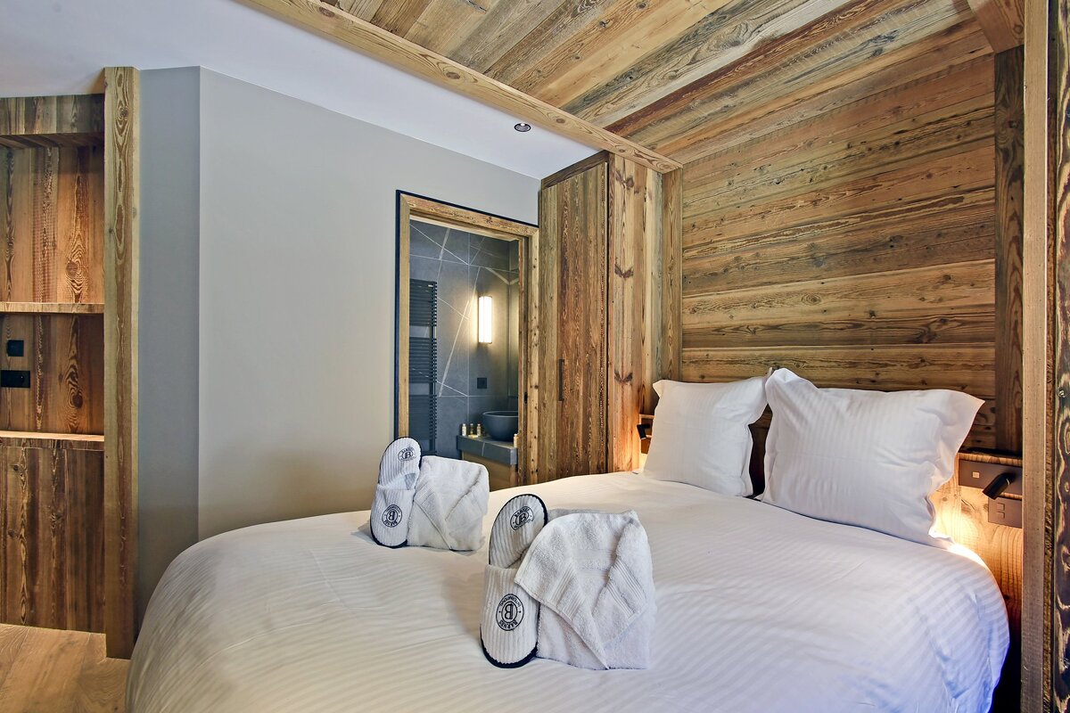 Cozy, rustic bedroom featuring wooden walls and ceiling, a white bed with pillows, slippers, and towels, with a bathroom visible beyond.