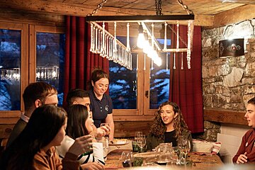 Friends enjoy a cozy dinner in a rustic chalet, served by staff under warm lighting. Dark windows suggest it's evening.