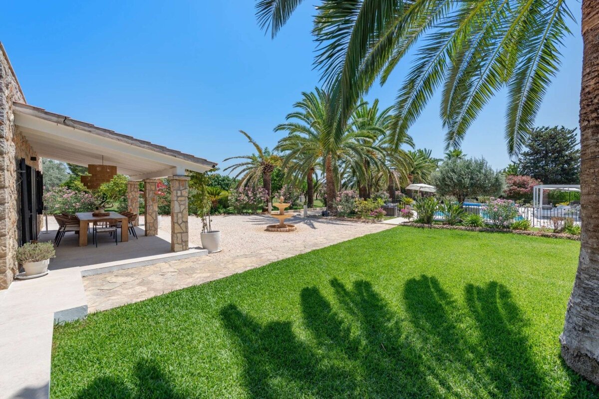 A bright, sunny outdoor scene featuring a stone patio with dining, a lush green lawn, many palm trees, a fountain, and a pool.