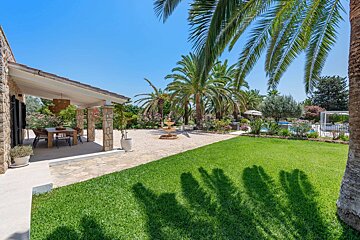 A spacious villa garden featuring a covered patio, manicured lawn, numerous palm trees, a fountain, and a pool area under a clear blue sky.