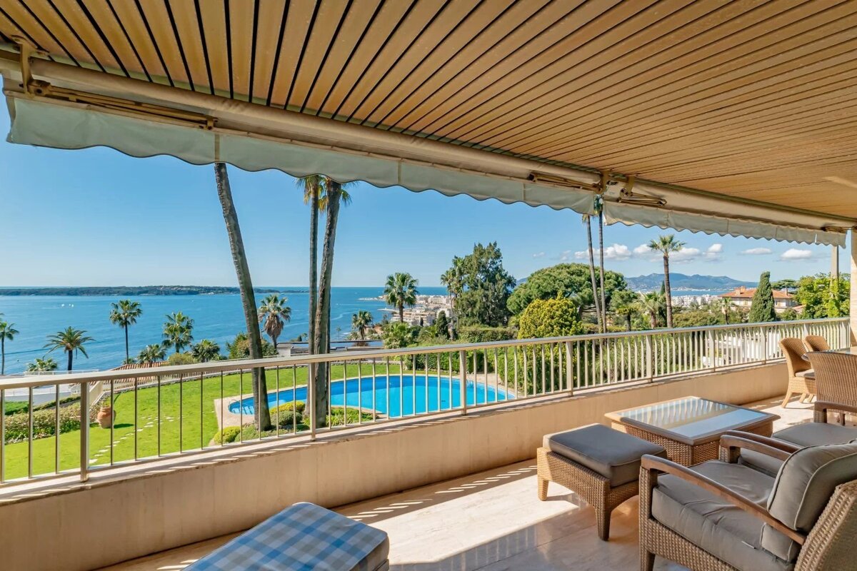 Sunny balcony with outdoor seating overlooking a swimming pool, palm trees, and a scenic blue ocean view.