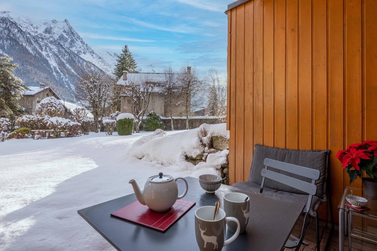 A cozy outdoor table with a teapot and mugs on a snowy balcony, offering picturesque views of a winter mountain village bathed in sunlight.