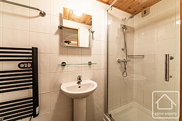 A bright bathroom with white tiled walls, a pedestal sink, mirror, glass-enclosed shower, black towel radiator, and a wooden ceiling.