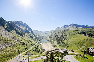 Sunny mountain valley with a deep blue lake, green slopes, village buildings, and ski lifts under a clear sky.