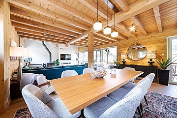 Warm, open-plan chalet dining & living area. Features a wooden table, modern grey chairs, and a comfy teal sofa, all with wood walls and beamed ceilings.