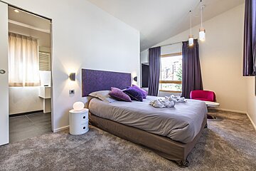 Modern bedroom featuring a bed with a purple headboard, grey bedding, and an open door leading to an en-suite bathroom.