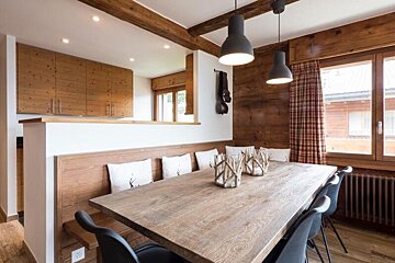Cozy rustic dining room featuring a long wooden table, built-in bench with pillows, black modern chairs, and warm wood-paneled walls.