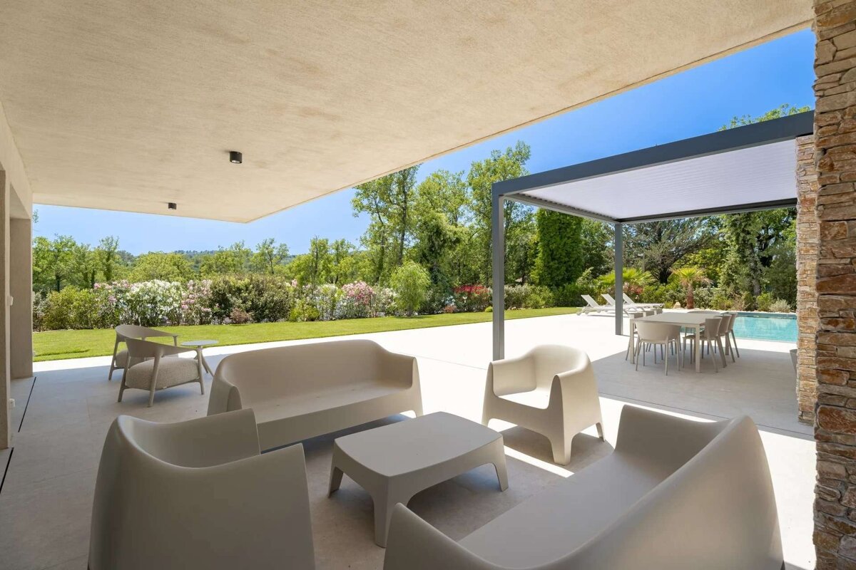 Modern outdoor living area with beige patio furniture under an overhang, looking out onto a lush garden, pool, and pergola.