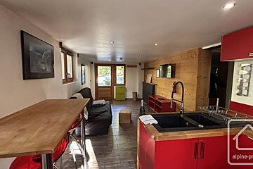 An open-plan living space with a red kitchen, wooden bar, couch, and TV on a wood-paneled wall. Double doors lead outside.