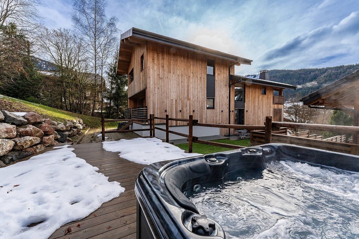 A bubbling hot tub on a snowy wooden deck outside a modern wooden chalet in the mountains.