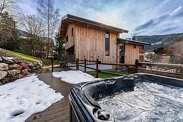 A bubbling hot tub on a snow-dusted wooden deck in front of a modern wooden chalet, with mountains and trees in the background.