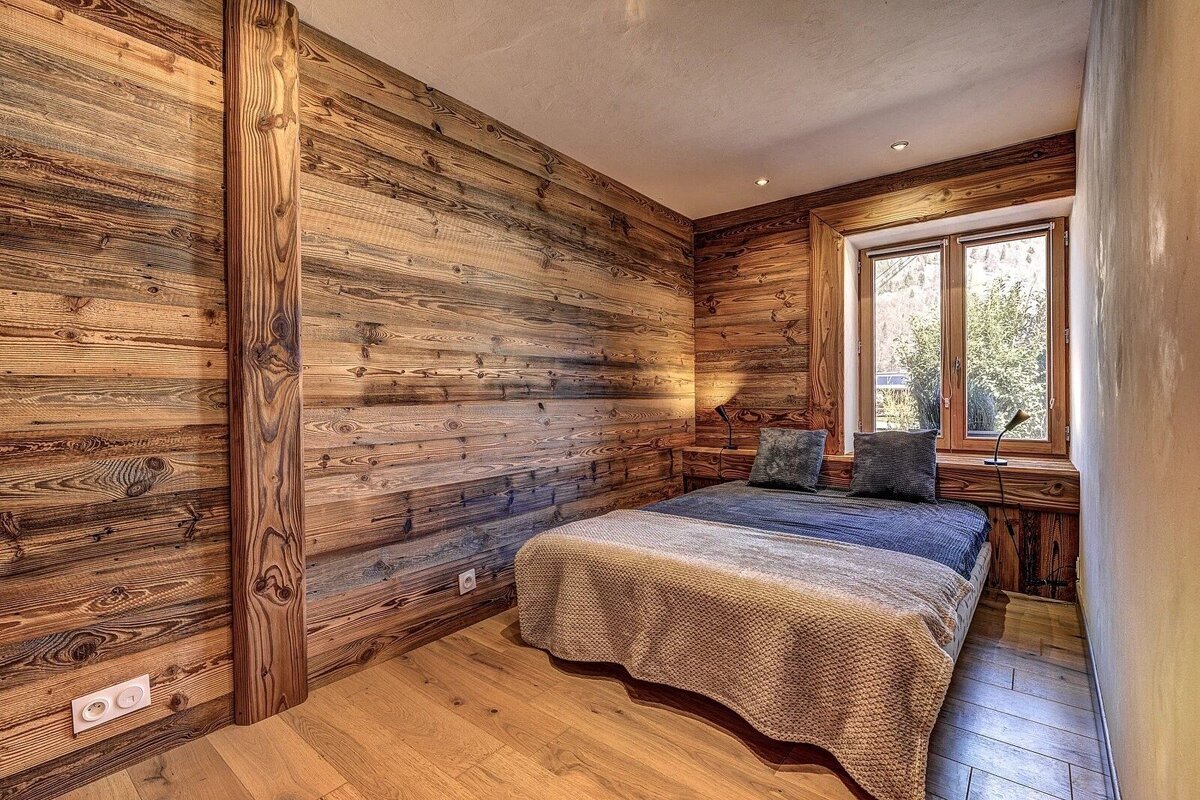 A cozy, rustic bedroom with warm wood-paneled walls and floor. It features a bed with pillows, two lamps, and a window letting in natural light.